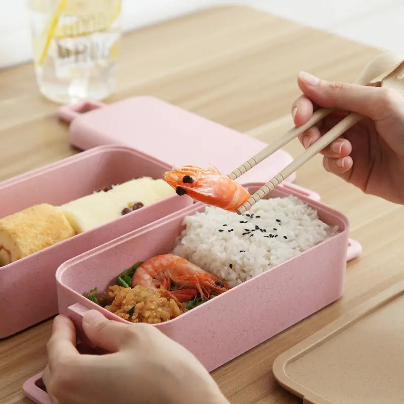 Pink bento box with food and a hand using chopsticks to pick up shrimp.